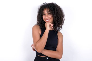 Optimistic Young African American woman wearing black T-shirt over white background keeps hands partly crossed and hand under chin, looks at camera with pleasure. Happy emotions concept.