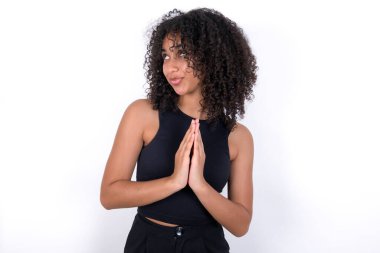 Charming cheerful Young African American woman wearing black T-shirt over white background making up plan in mind holding hands together, setting up an idea. Look askance