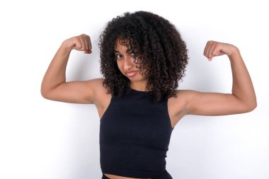 Beyaz arka plan üzerine siyah tişört giyen genç Afrikalı Amerikalı kadın gururla gülümseyen kol kaslarını gösteriyor. Fitness konsepti.