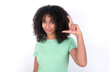 Young African American woman wearing green T-shirt over white background smiling and confident gesturing with hand doing small size sign with fingers looking and the camera. Measure concept