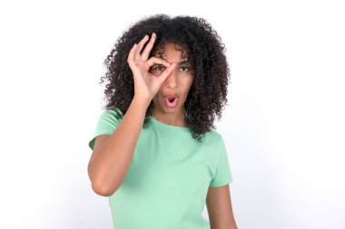 Young African American woman wearing green T-shirt over white background doing ok gesture shocked with surprised face, eye looking through fingers. Unbelieving expression.