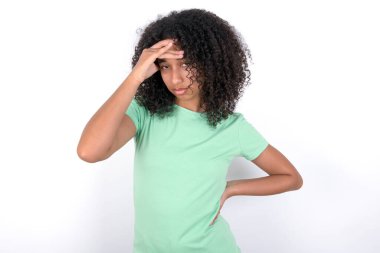 Young African American woman wearing green T-shirt over white background having problems, worried and stressed holds hand on forehead.