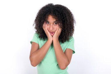 Stupefied Young African American woman wearing green T-shirt over white background expresses excitement and thrill, keeps jaw dropped, hands on cheeks, has eyes popped out