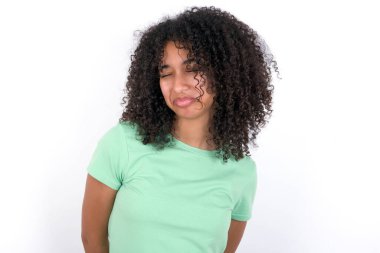 Dismal gloomy rejected Young African American woman wearing green T-shirt over white background has problems and difficulties, curves lower lip and closes eyes in despair, being in depression