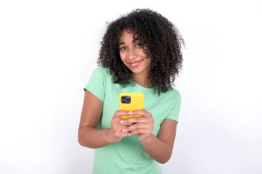 Beyaz arka planda yeşil tişört giyen, elinde akıllı telefon tutan heyecanlı Afrikalı Amerikalı kadın iyi haberler aldıktan sonra kameraya hayret ediyor..