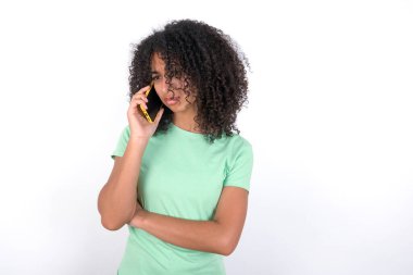 Sad Young African American woman wearing green T-shirt over white background talking on smartphone. Communication concept.