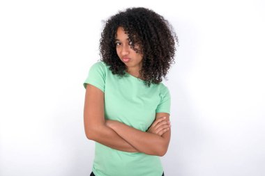 Gloomy dissatisfied Young African American woman wearing green T-shirt over white background looks with miserable expression at camera from under forehead, makes unhappy grimace