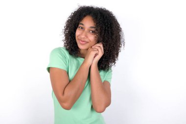 Charming serious Young African American woman wearing green T-shirt over white background keeps hands near face smiles tenderly at camera