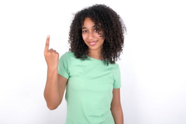 Young African American woman wearing green T-shirt over white background smiling and looking friendly, showing number one or first with hand forward, counting down