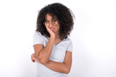 Sad lonely Young African American woman wearing grey T-shirt over white background touches cheek with hand bites lower lip and gazes with displeasure. Bad emotions