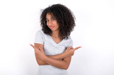 Young African American woman wearing grey T-shirt over white background crosses arms and points at different sides hesitates between two items or variants. Needs help with decision