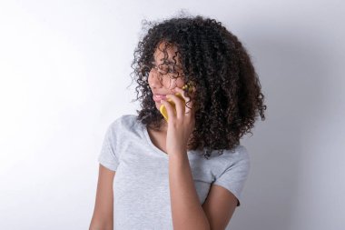 Pleasant looking happy Young African American woman wearing grey T-shirt over white background has nice telephone conversation and looks aside, has nice mood and smiles positively while talks via cell phone