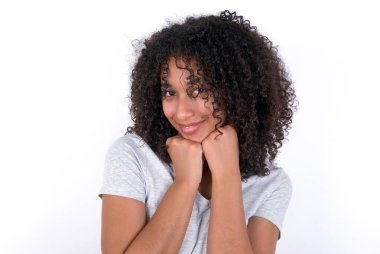 Satisfied Young African American woman wearing grey T-shirt over white background touches chin with both hands, smiles pleasantly, rejoices good day with lover