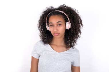 Serious displeased Young African American woman wearing grey T-shirt over white background looks puzzled at camera being angry wears stereo headphones listens music while walking at street