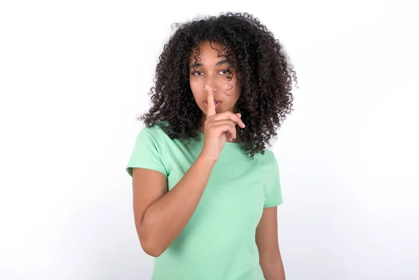 白い背景に緑色のTシャツを着た若いアフリカ系アメリカ人女性は 沈黙のジェスチャーを行い 唇の上に指を保持します 沈黙と秘密の概念 — ストック写真