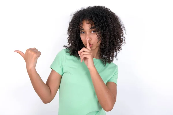 白い背景の上に緑色のTシャツを着た若いアフリカ系アメリカ人女性は 唇に手を指差して静かになるように求めています 沈黙と秘密の概念 — ストック写真