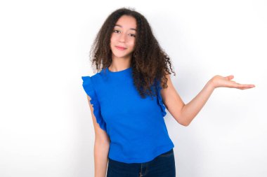 Afro saçlı genç kız beyaz arka planda mavi tişört giyiyor neşeli bir şekilde gülümsüyor ve avuç içi kameraya bakıyor..