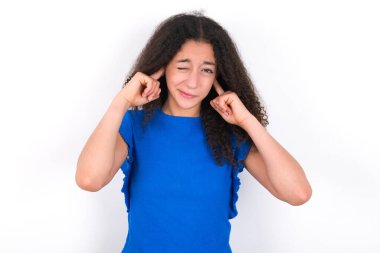 Afro saçlı genç kız, yüksek sesli müzik sesi için beyaz arka plan üzerine mavi tişört giyip kulaklarını parmaklarıyla kapatıyor. Sağır kavramı.