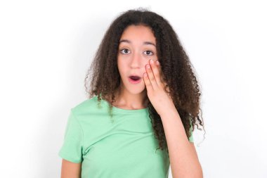 Şok olmuş, afro saçlı, yeşil arka planda beyaz tişört giyen genç bir kız çok şaşırmış görünüyor. Beklenmedik haberler, yüz ifadeleri konsepti....