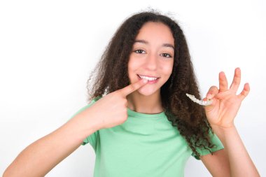 Teenager girl with afro hairstyle wearing white T-shirt over green background holding an invisible aligner and pointing to her perfect straight teeth. Dental healthcare and confidence concept.