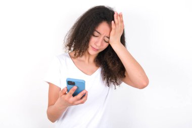 Upset depressed Teenager girl with afro hairstyle wearing white T-shirt over white background makes face palm as forgot about something important holds mobile phone expresses sorrow and regret blames