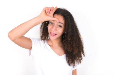 Beyaz arka planda beyaz tişörtlü, afro saçlı komik genç kız, dillerini çıkarıp suratını buruşturan birine ezik jestler yapıyor..