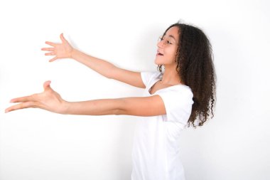 Funny astonished Teenager girl with afro hairstyle wearing white T-shirt over white background look empty space with arms opened ready to catch something.