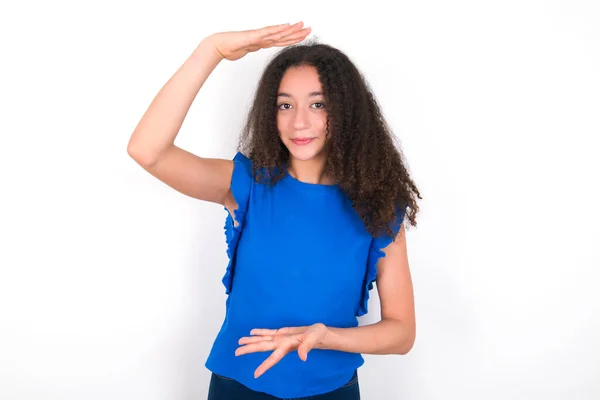 青のTシャツを身に着けているアフロの髪型を持つ10代の女の子は 手が大きく 大きなサイズのサインを示すと白い背景ジェスチャー上にTシャツ メジャーシンボル カメラを見て微笑んだ — ストック写真