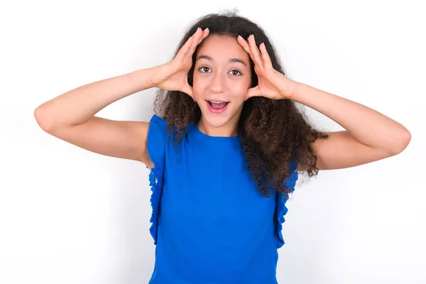 怖い表情で白い背景に青いTシャツを着てアフロの髪型を持つ10代の女の子は 頭の上に手を維持し 顎を落とした 素晴らしい表現をしています Omgの概念 — ストック写真