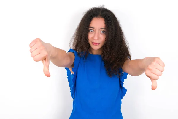 白い背景に青いTシャツを着たアフロの髪型をした10代の少女が両手で親指を下に見せて動揺している コンセプトが嫌い — ストック写真