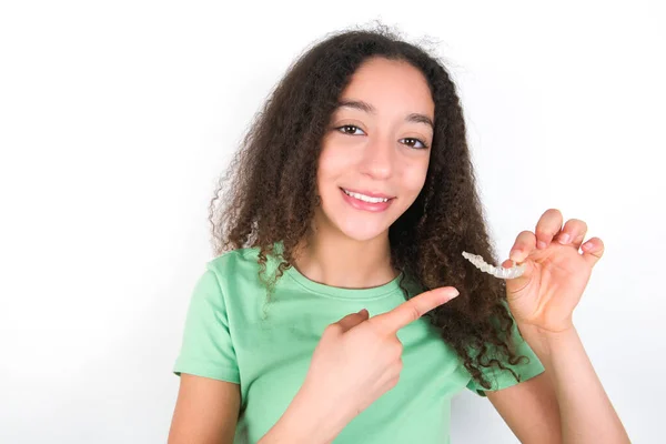 Afro saçlı, yeşil arka planda beyaz tişört giyen genç kız görünmez bir hizalama aleti tutuyor ve onu işaret ediyor. Diş sağlığı ve güven kavramı.
