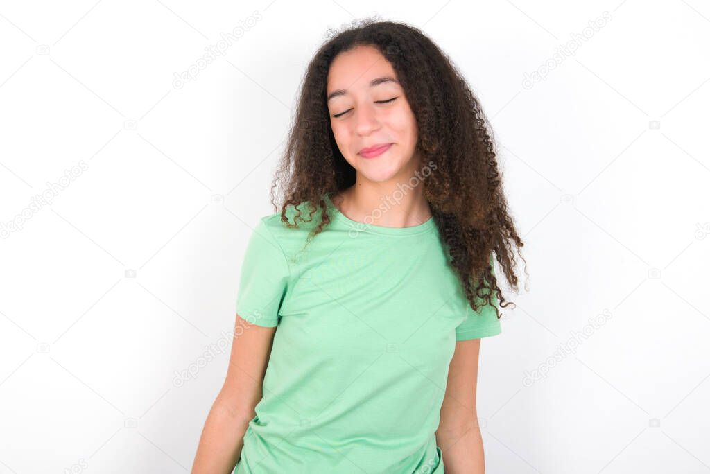 Positive Teenager girl with afro hairstyle wearing white T-shirt over green background with overjoyed expression closes eyes and laughs shows white perfect teeth