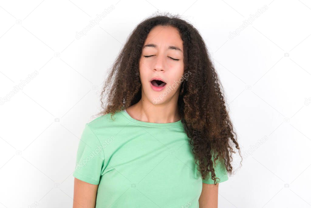 Teenager girl with afro hairstyle wearing white T-shirt over green background yawns with opened mouth stands. Daily morning routine