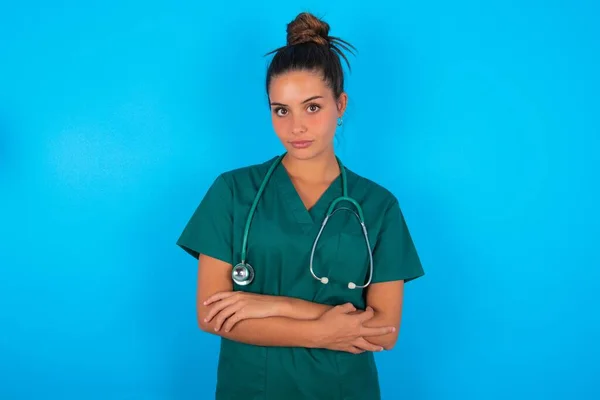 青の背景に緑の医療制服を身に着けている深刻な情熱的な美しいヒスパニック系の医師の女性はクールな自信起業家クロス手のように感じる — ストック写真