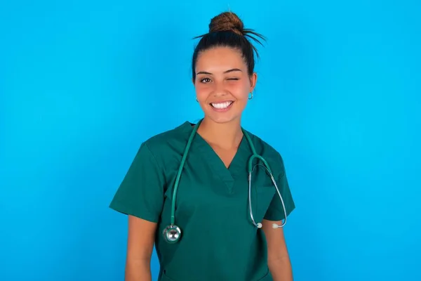 非常に美しいですヒスパニック系の医師の女性身に着けています緑の医療制服上の青の背景笑顔幸せに 遊び心のある方法でカメラで点滅 あなたといちゃつく — ストック写真