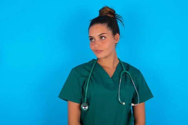 青い背景の上に緑の制服を着た美しいヒスパニック系の医者の女性が空の空間に思いやりを脇に見て — ストック写真