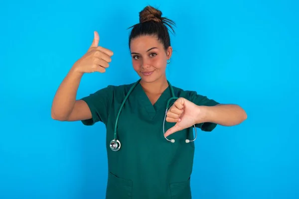 青い背景の上に緑色の制服を着た美しいヒスパニック系の医者の女性が親指を立てて難しい選択の概念 — ストック写真