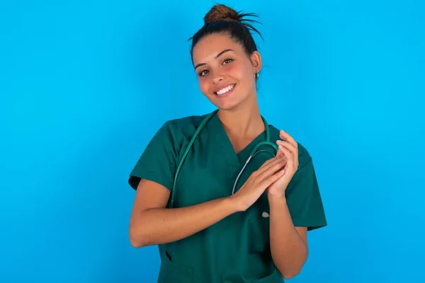 青い背景に緑色の制服を着た美しいヒスパニック系の医師の女性が拍手と幸せと喜びに満ちた笑顔の誇り高い手を一緒に — ストック写真