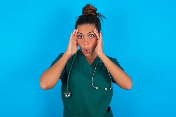 恐怖の表情で青の背景に緑の医療制服を着た美しいヒスパニック系の医師の女性は 頭の上に手を維持し 素晴らしい表現を持ってドロップされます Omgの概念 — ストック写真