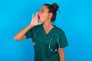Mavi arka planda yeşil üniforma giyen güzel İspanyol doktor kadın el ve ağızla bağırarak ve yüksek sesle bağırıyordu. İletişim konsepti.
