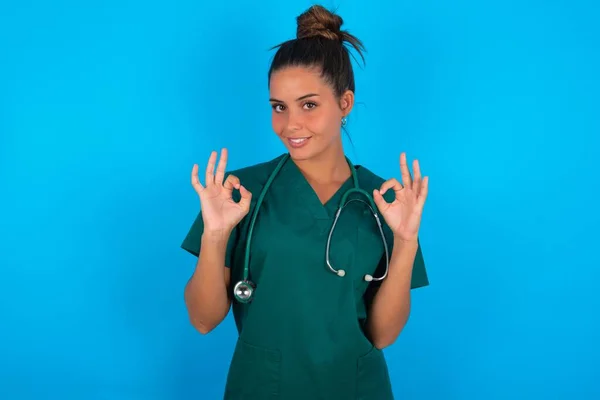 青い背景に緑色の制服を着た美しいヒスパニック系の医師の女性は Okサインで指で両手を示しています コンセプトの承認または推奨 — ストック写真