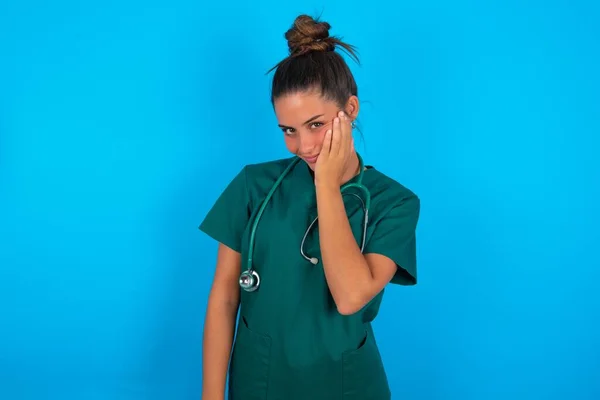 非常に退屈美しいですヒスパニック系の医師の女性は 別の交差手でそれをサポートしながら 頬に手を保持青い背景に緑の医療制服を身に着けています 疲れていると病気に見えます — ストック写真