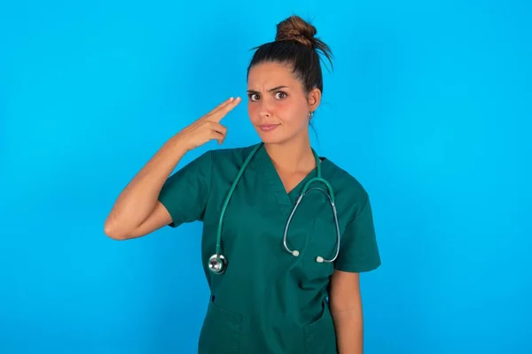 青い背景に緑の制服を着た不幸な美しいヒスパニック系の医師の女性は自殺ジェスチャーを行い 手で銃を模倣 曲線の唇 寺院で2本の指を保持 — ストック写真