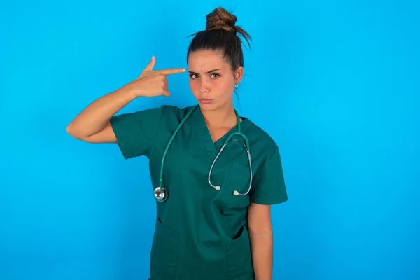 青い背景の上に緑の医療制服を着た不幸な美しいヒスパニック系の医師の女性は自殺ジェスチャーをし 手で銃を模倣し 曲線の唇は寺院で2本の指を保持し すべての疲れている — ストック写真