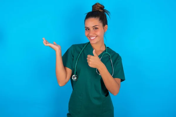 青い背景の上に緑の制服を着た美しいヒスパニック系の医師の女性ヤシの手を表示し 親指でOkジェスチャーを行います 笑顔幸せと陽気な — ストック写真