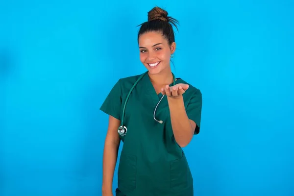 青い背景に緑色の制服を着た美しいヒスパニック系の医師の女性が笑顔明るい提供ヤシの手援助と受け入れを与えます — ストック写真
