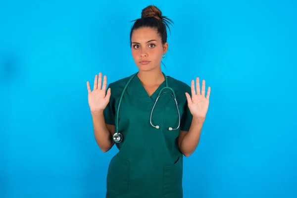 青の背景の上に緑の医療制服を着て深刻な美しいパニック医師の女性は カメラに向かって手のひらを引くと ジェスチャーを停止し あなたの感情を制御するために尋ねると緊張してはいけません — ストック写真