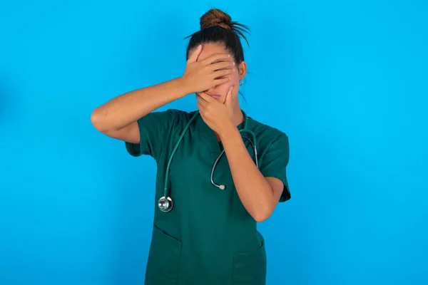 青い背景に緑の制服を着た美しいヒスパニック系の医師の女性手で目と口をカバー 驚きとショックを受けました 感情を隠す — ストック写真