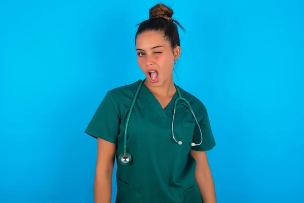 青い背景の上に緑の制服を着た美しいヒスパニック系の医師の女性がセクシーな表情でカメラを見てウィンク 陽気で幸せな顔 — ストック写真