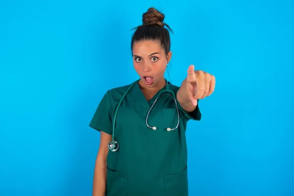 青い背景の上に緑の制服を着た美しいヒスパニック系の医師の女性指で先に驚いて 口を開けて驚くような表情を指し 正面に何か — ストック写真